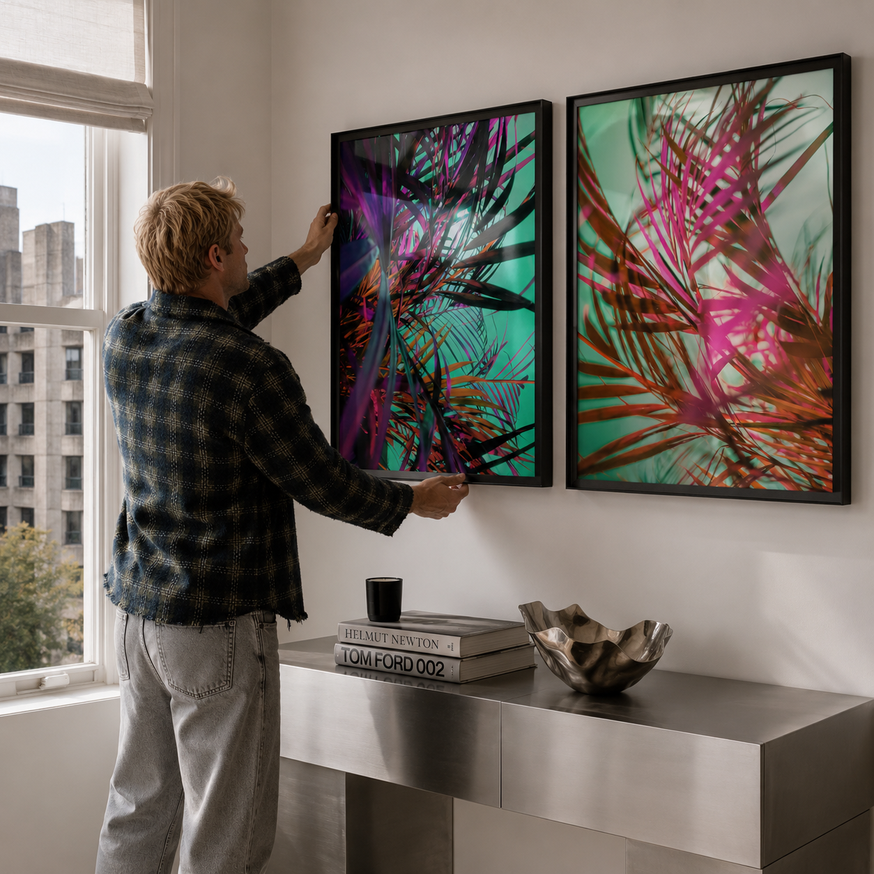 A man in a plaid shirt hangs two framed abstract botanical wall art prints on a white wall above a modern console table in a bright room.