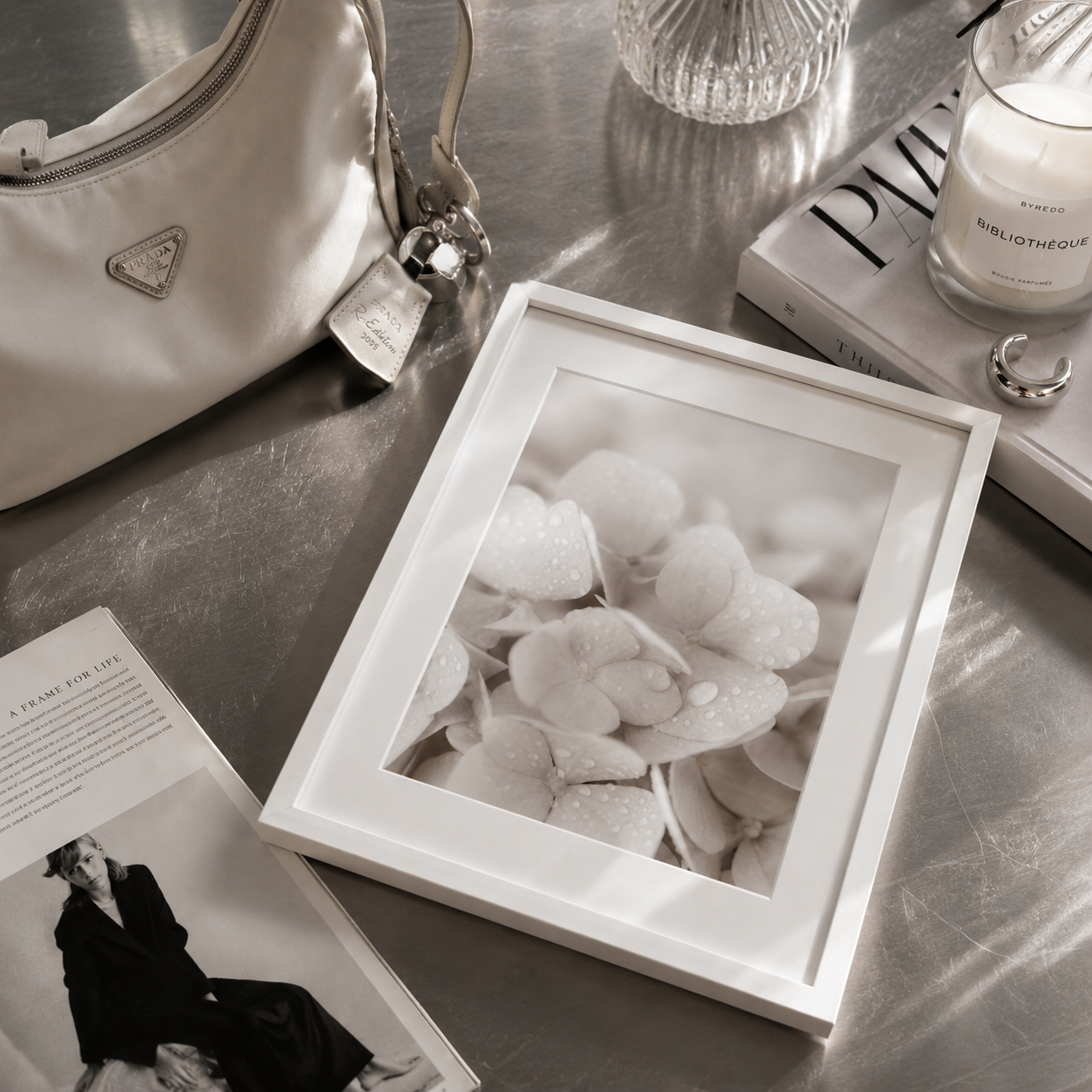 A white square picture frame displaying a black and white photograph of water droplets on flowers, styled on a metallic surface with a cream handbag, a candle, and a magazine nearby.