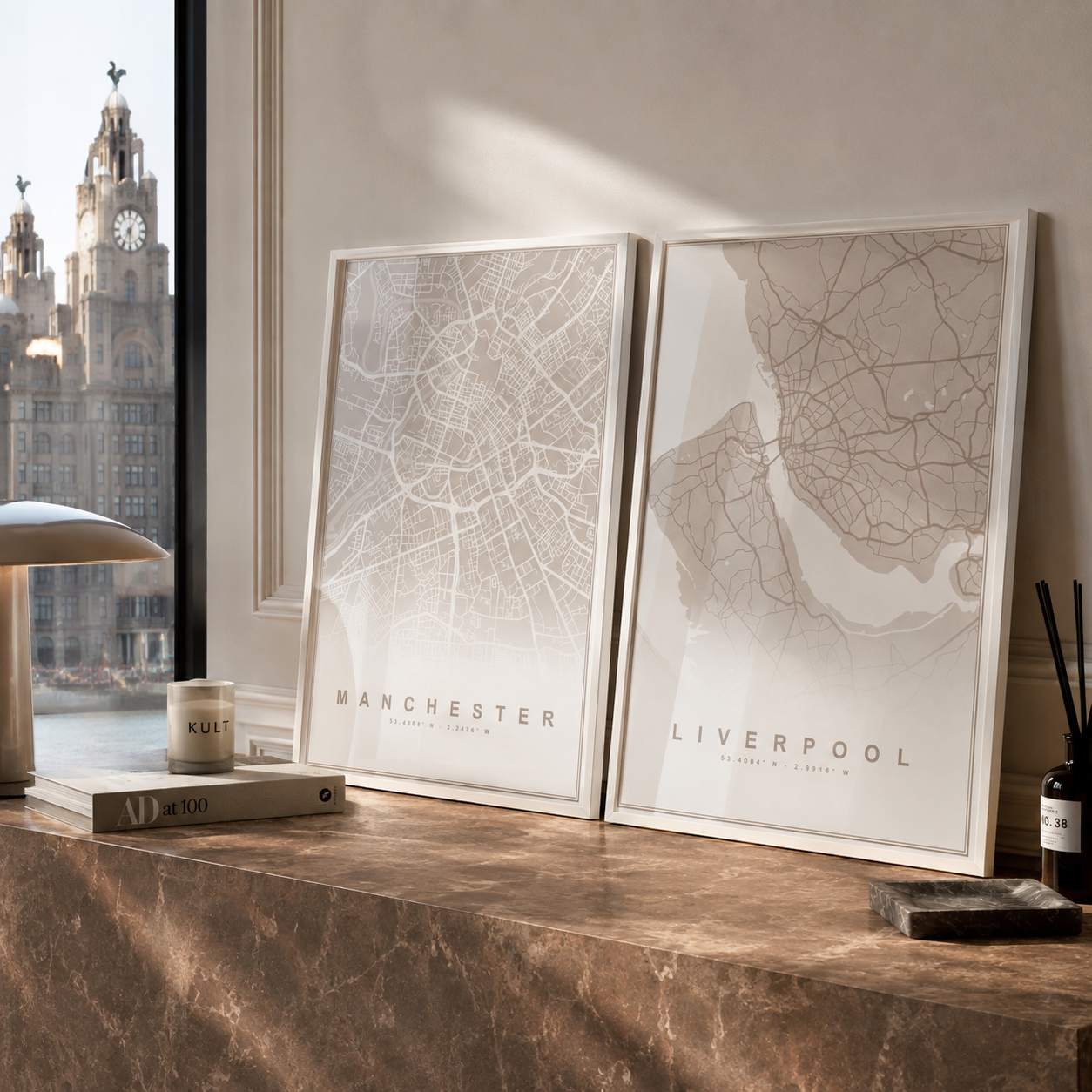 Two framed city map prints leaning on a marble console table in a styled interior. The left print is labeled Manchester and the right print is labeled Liverpool.
