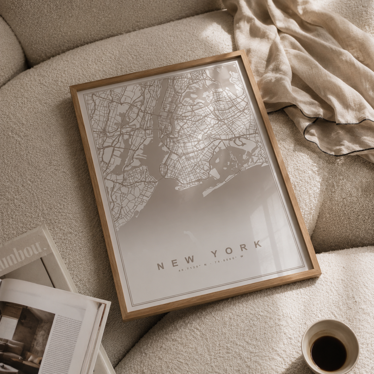 A framed New York City map art print with a light wood frame, styled on a beige textured sofa with a linen throw blanket, an open magazine, and a cup of coffee nearby.