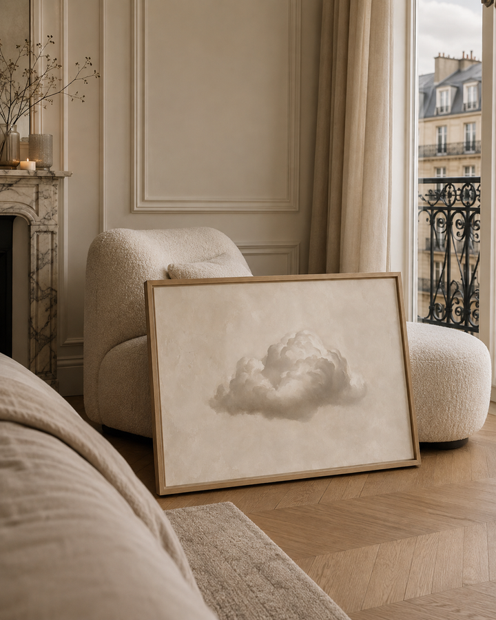 A framed art print featuring a soft gray cloud illustration on a textured cream background, styled in a cozy, elegant room next to a window with Parisian buildings outside.