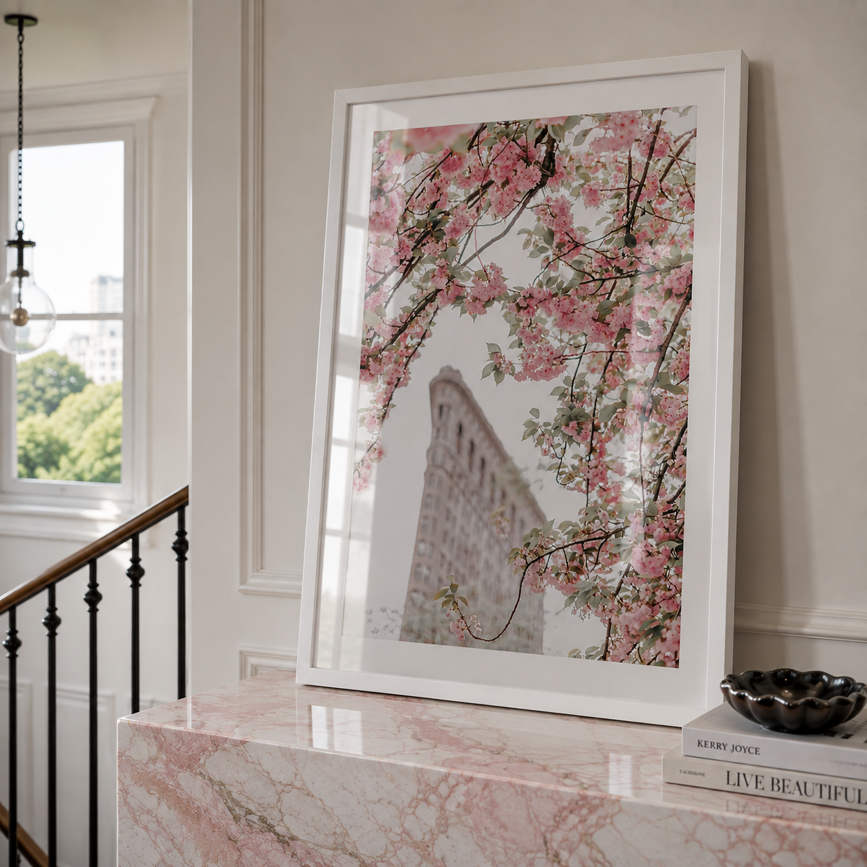 A framed art print featuring pink cherry blossoms and a building facade, displayed on a pink marble console table in a styled interior setting.
