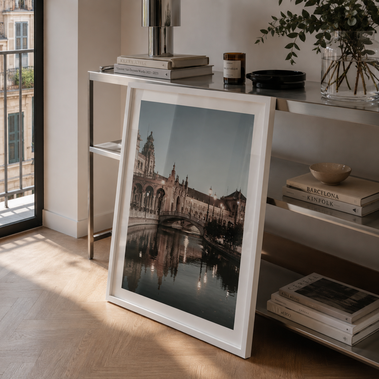 A framed black and white architectural art print leaning against a modern glass console table in a bright, sunlit room with wood floors and shelves.