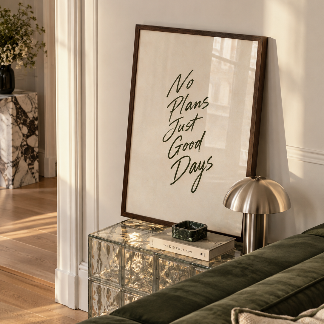 A framed wall art print with the phrase 'No Plans Just Good Days' in black script on a warm off-white background, leaning against a white wall in a styled living room next to a green velvet sofa and a metallic table lamp.