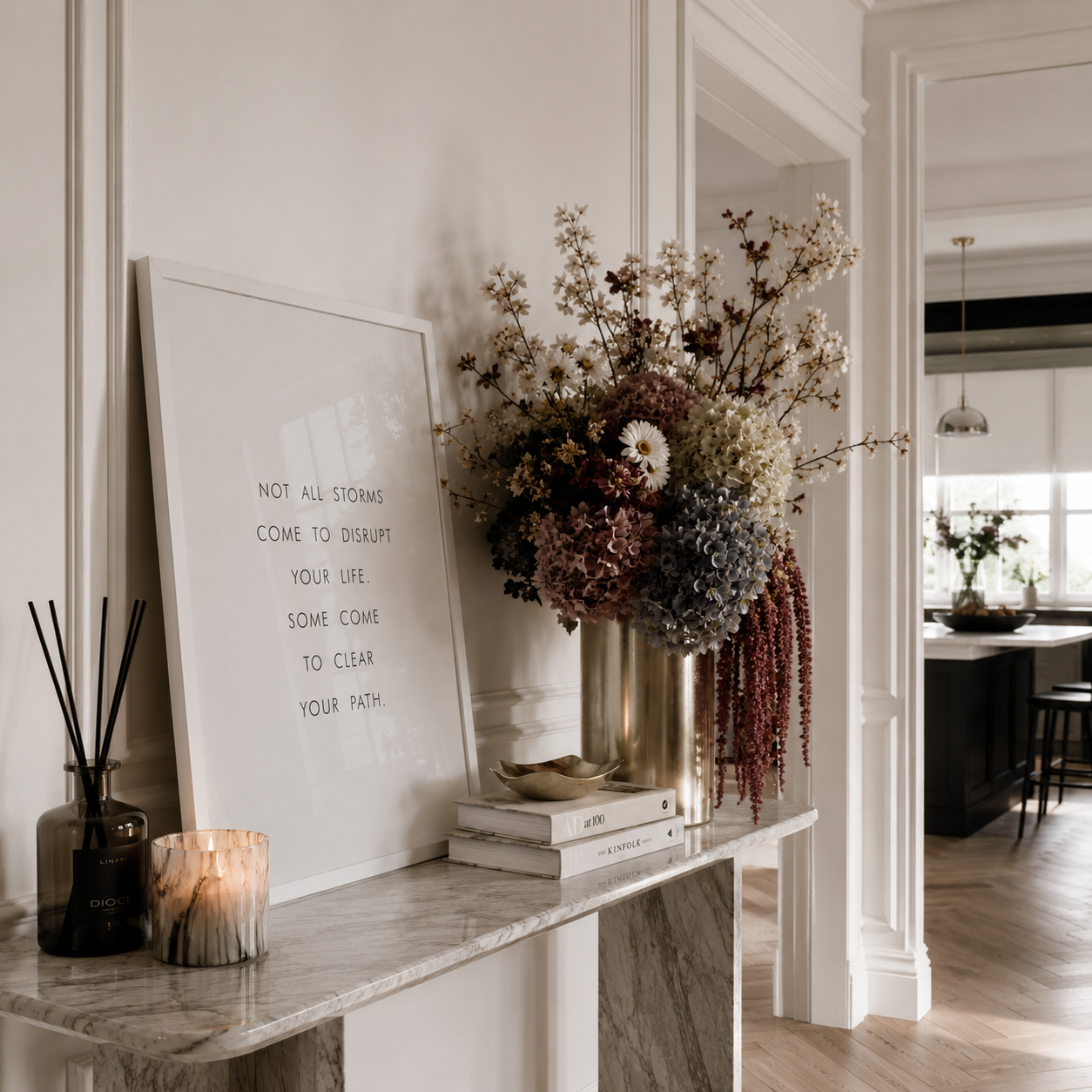 A styled interior scene with a white framed art print leaning on a marble console table, next to a large dried floral arrangement in a metallic vase, a candle, and a reed diffuser. The room has white walls, molding, and a visible kitchen in the background.