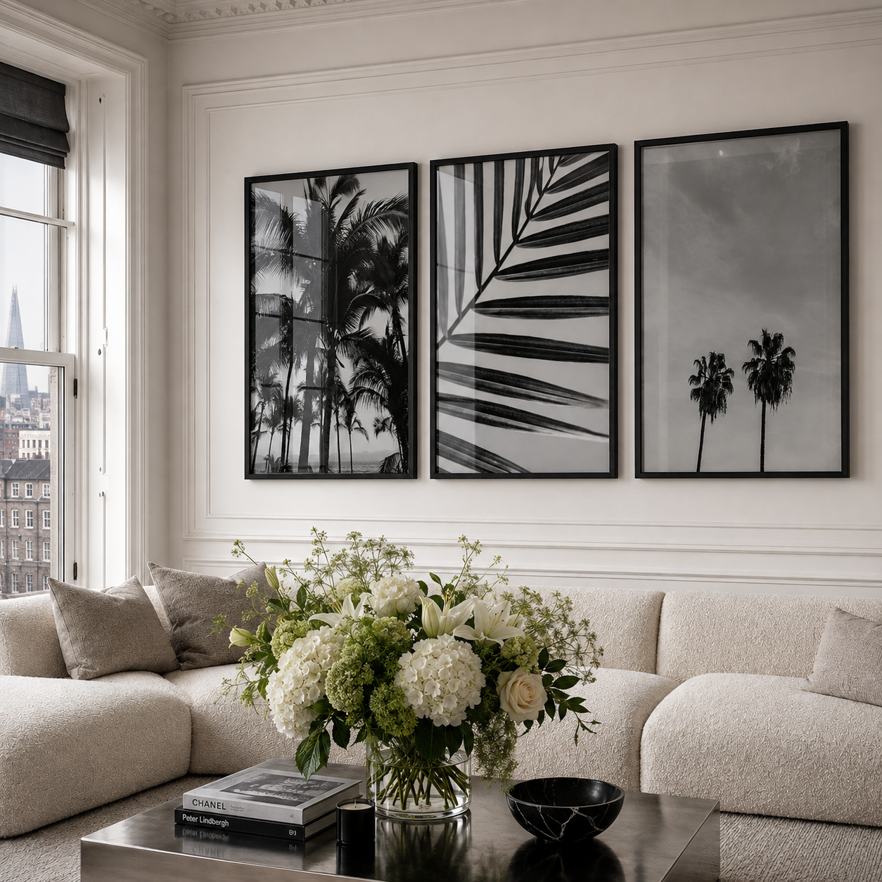 Three framed black and white botanical and palm tree wall art prints hang above a cream boucle sofa in a bright living room with a floral arrangement and coffee table.