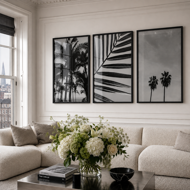 Three framed black and white botanical and palm tree wall art prints hang above a cream boucle sofa in a bright living room with a floral arrangement and coffee table.