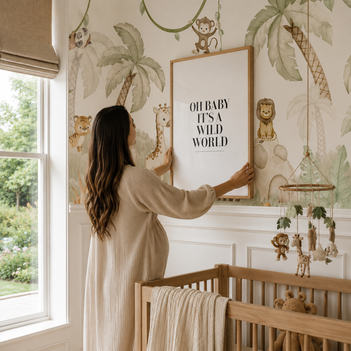 A pregnant woman in a beige robe hangs a framed nursery print on a tropical safari-themed wallpapered wall next to a wooden crib.