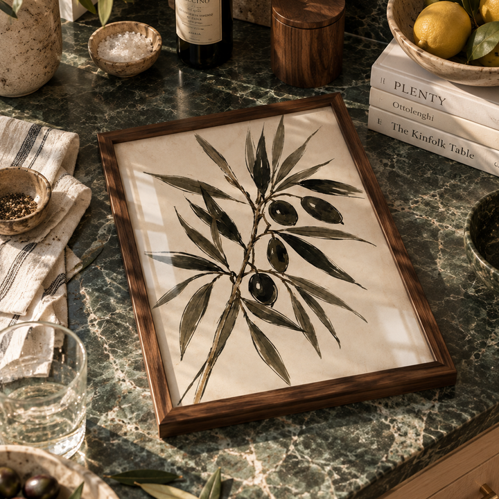 A rectangular wooden-framed art print featuring a botanical illustration of an olive branch with dark olives, displayed on a green marble countertop surrounded by lifestyle props including olives, a glass of water, books, and a bottle.