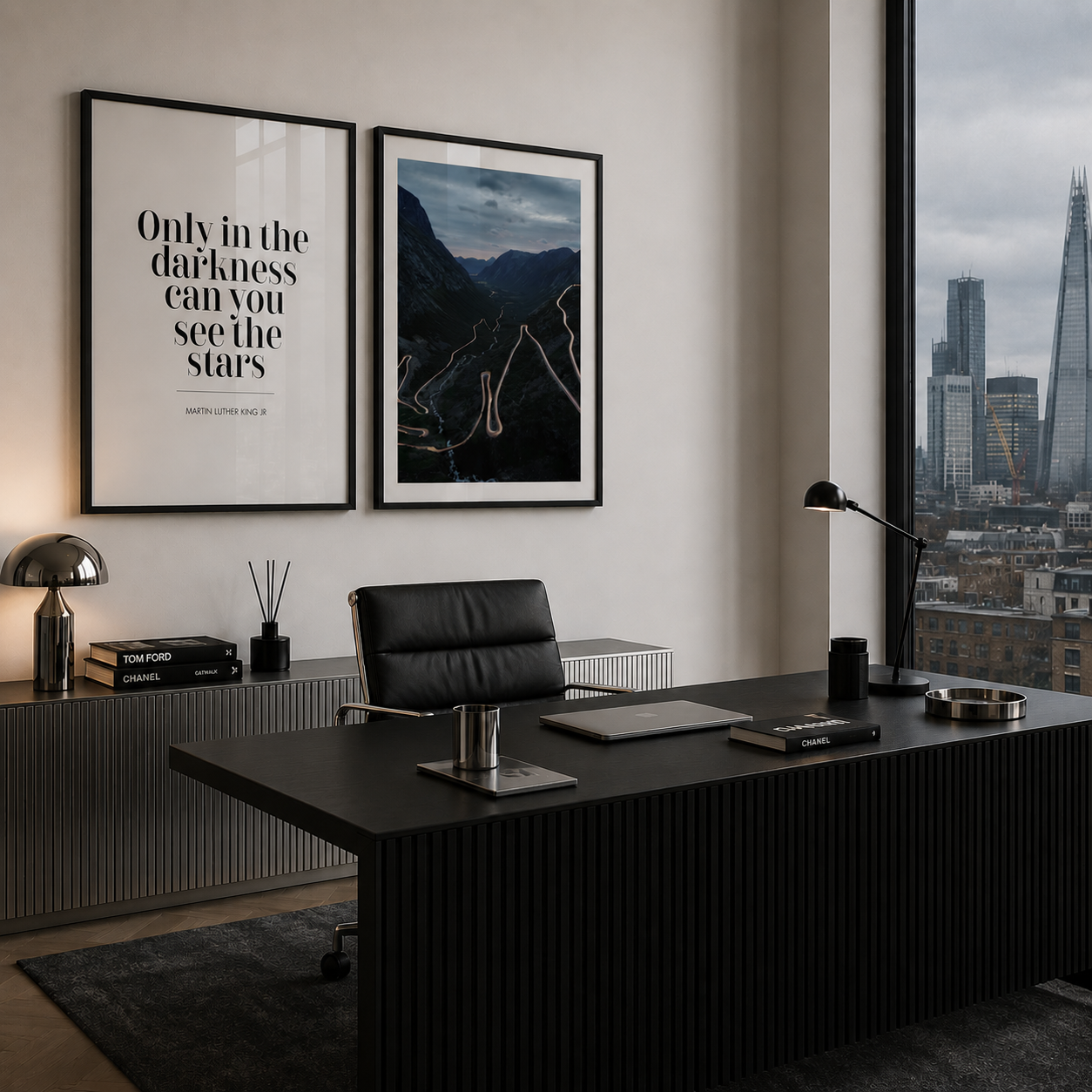 A modern office interior featuring two framed wall art prints hanging above a black desk. The left print has black text on a white background reading 'Only in the darkness can you see the stars', and the right print shows a dark mountain landscape.
