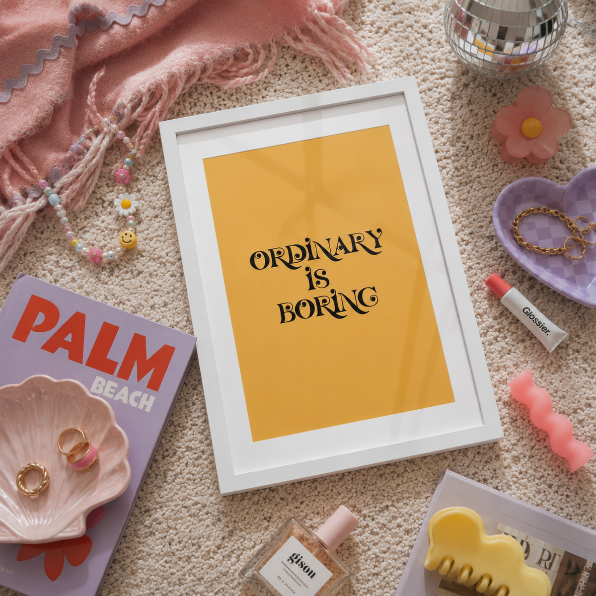 A framed typographic art print reading 'ORDINARY IS BORING' in black script on a mustard yellow background, styled in a flat lay on a beige carpet with pink accessories, jewelry, a magazine, and small decor items around it.