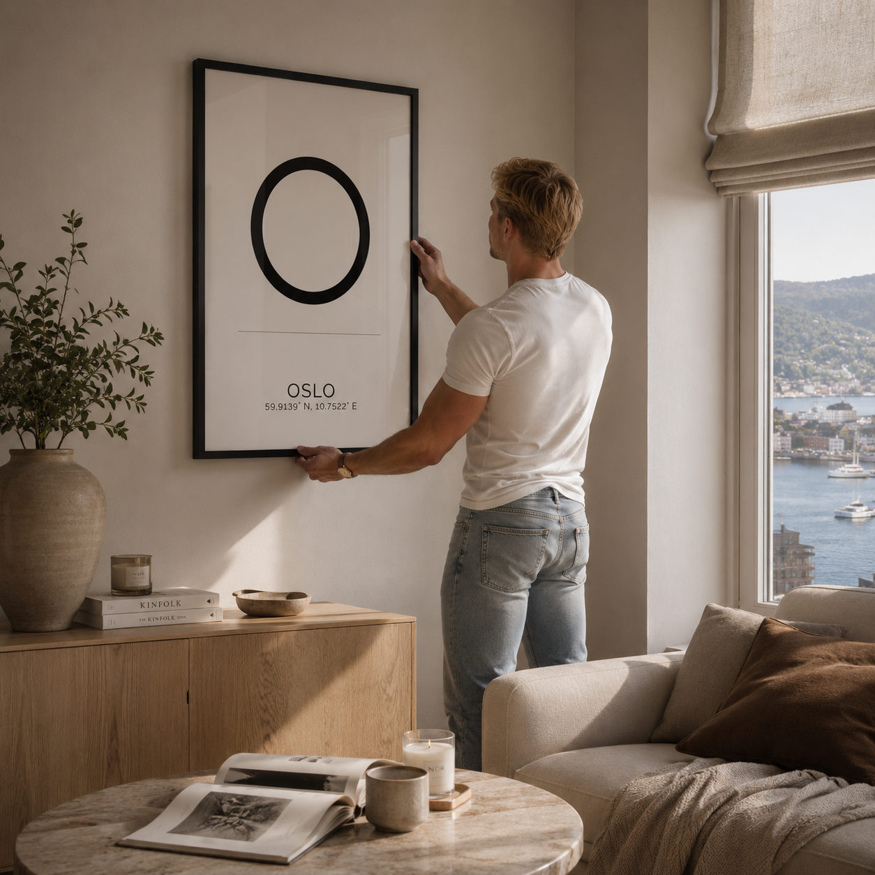 A man in a white t-shirt and light blue jeans hangs a minimalist black and white framed art print on a beige wall in a modern, sunlit living room. The print features a large black circle on a white background with the text 'OSLO' and smaller text below.