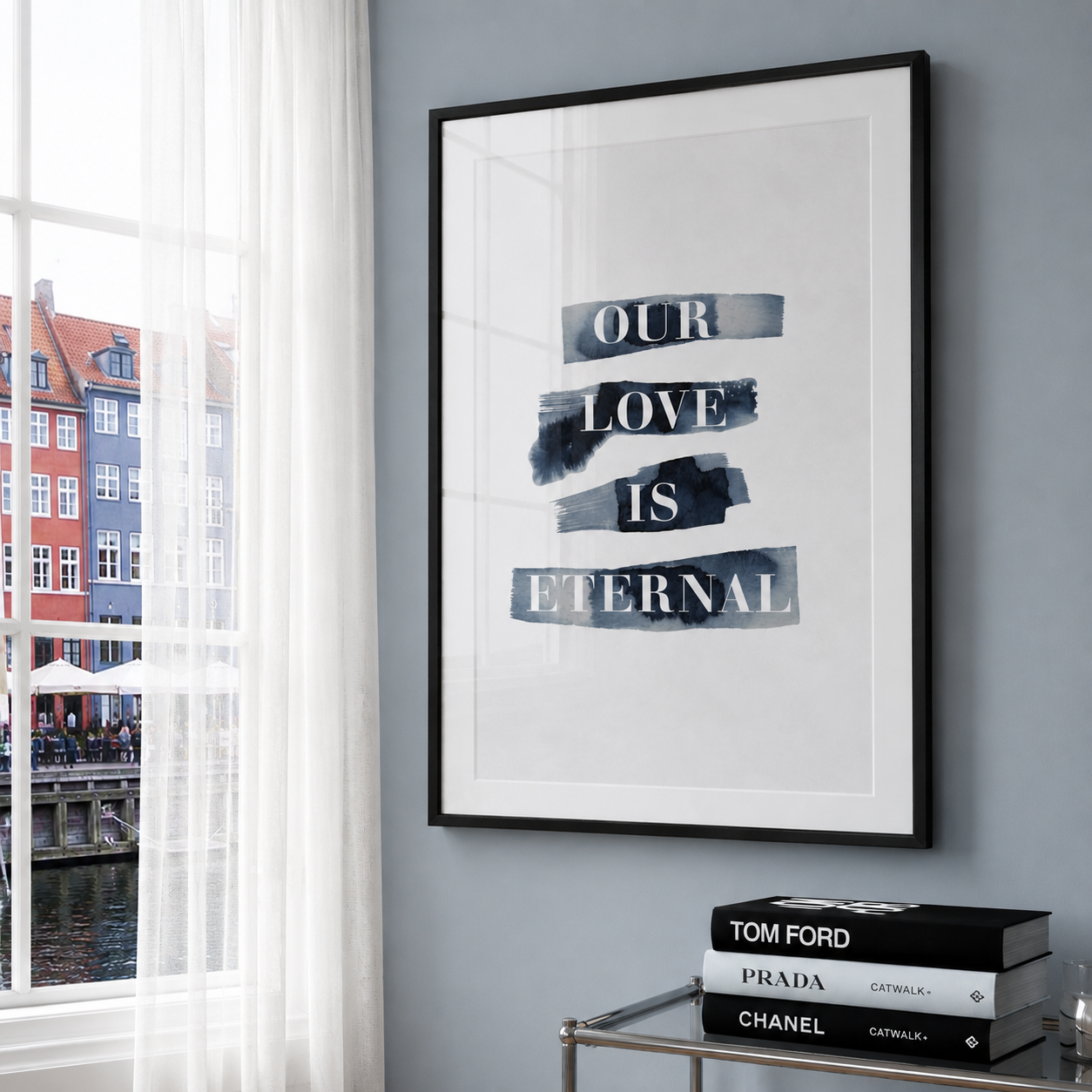 A framed wall art print with the text 'OUR LOVE IS ETERNAL' in white serif letters on layered dark blue-gray brushstroke bands, displayed on a light gray wall beside a window with white curtains and a small stack of books below.