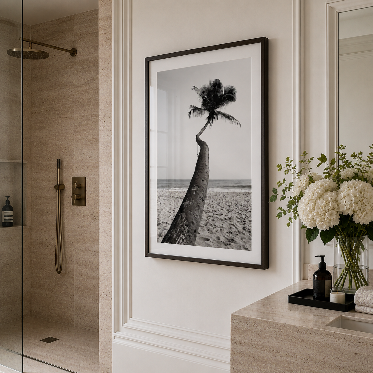 A framed black and white beach photograph featuring a leaning palm tree, displayed on a white wall in a modern bathroom setting with a stone shower and a marble vanity with white flowers.