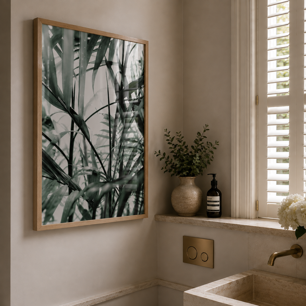 A framed botanical art print with green palm leaves on a light gray background, styled in a modern bathroom next to a window with white shutters and a stone sink.