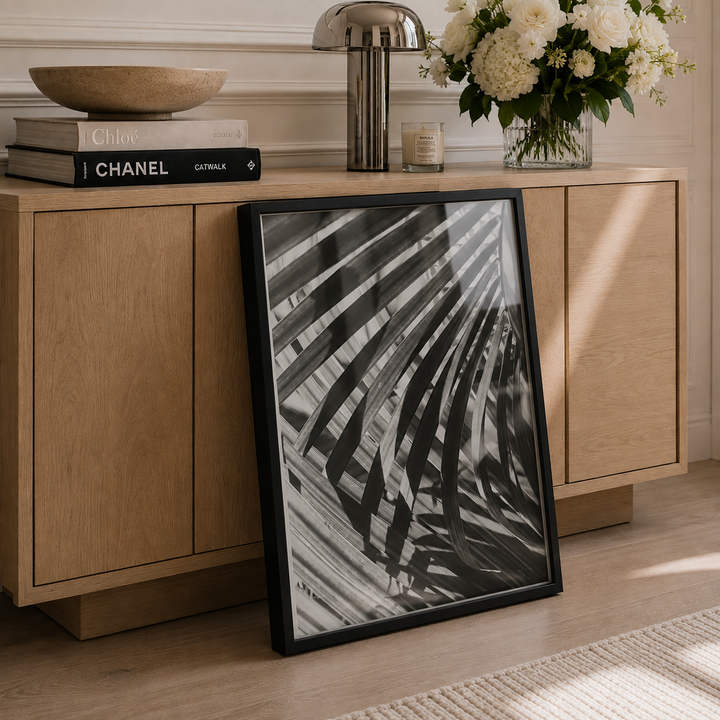 A black-framed black and white botanical art print leaning against a light wood sideboard in a styled room setting.