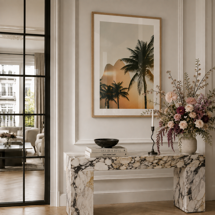 A framed art print featuring a tropical sunset with palm trees, displayed on a white paneled wall above a marble console table in a beautifully styled room.