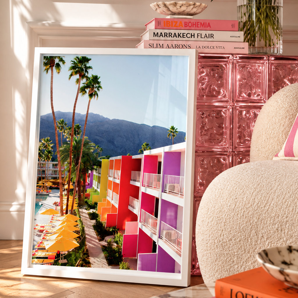 A framed art print leaning against a white wall in a stylishly decorated room. The print features a colorful mid-century modern motel scene with palm trees and mountains.