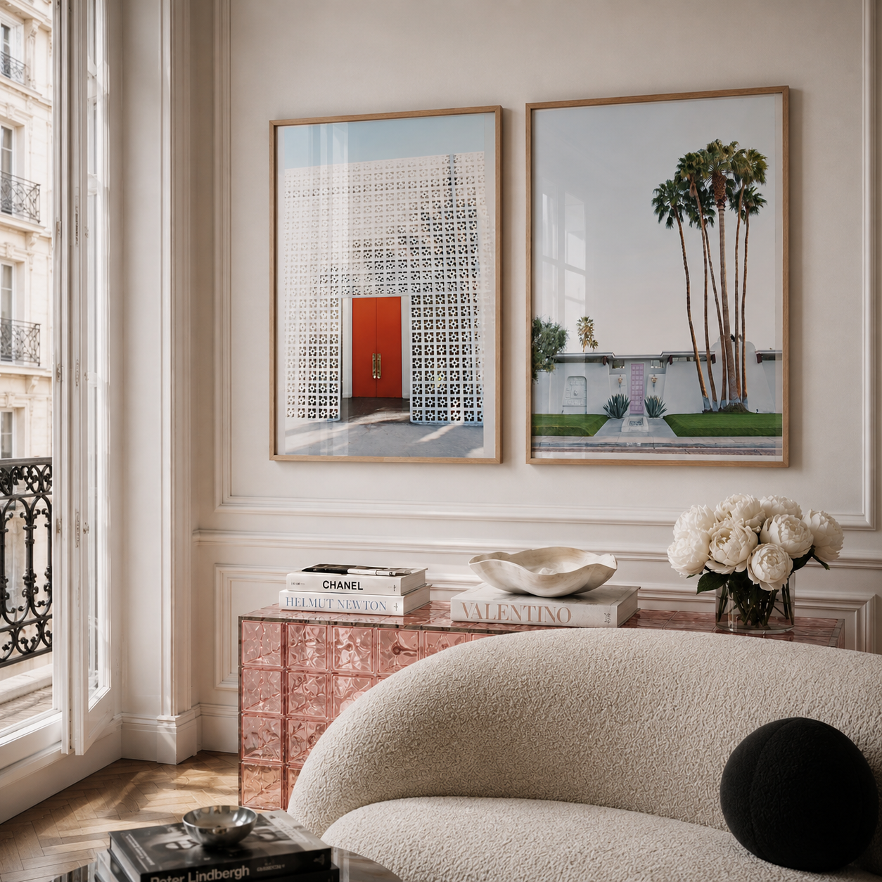 Two framed art prints hanging on a cream-colored wall in a stylishly decorated room. The left print features a modern white lattice structure with a bold red door, and the right print shows tall palm trees in a driveway.