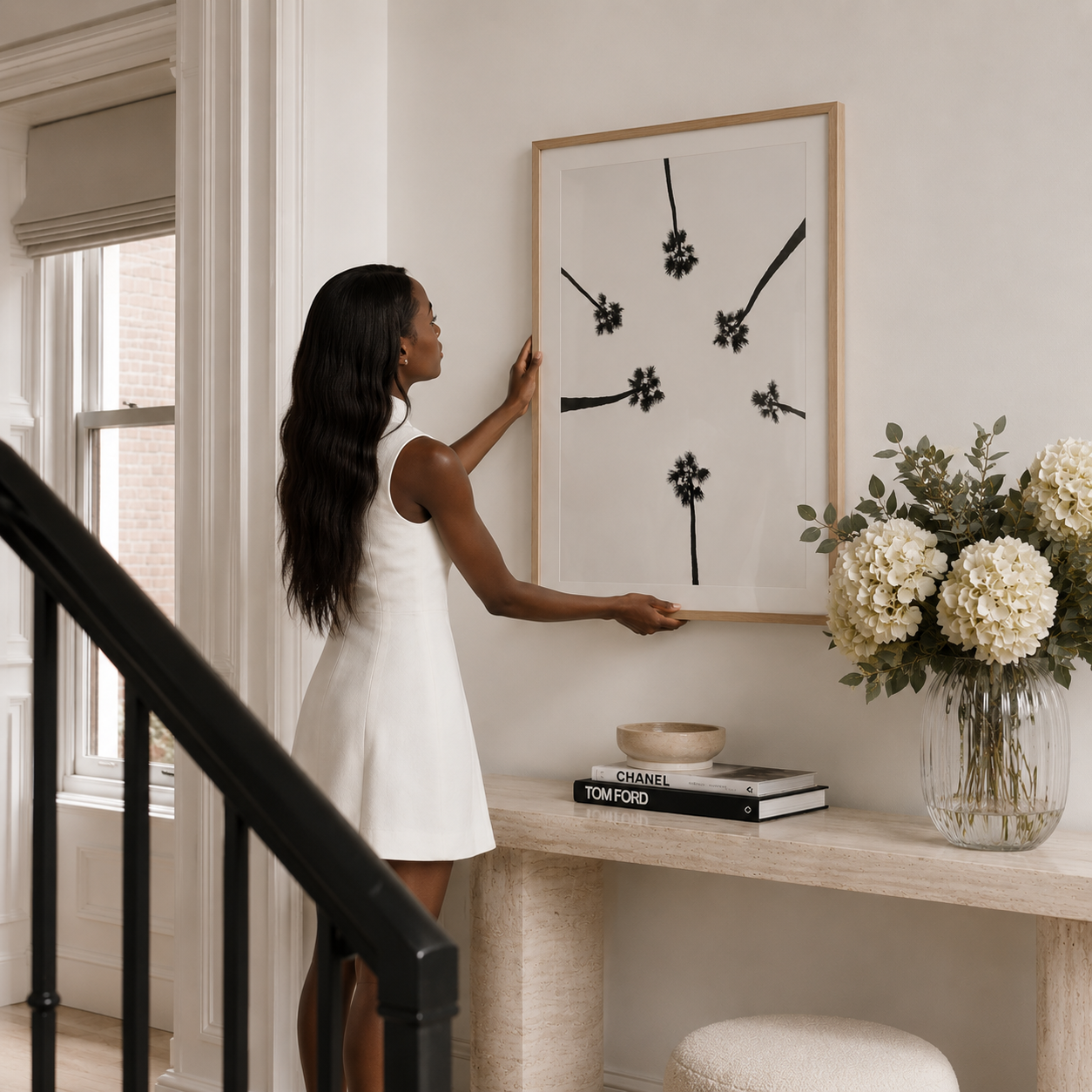 A woman in a white sleeveless dress hangs a large framed black-and-white abstract floral artwork on a cream wall above a console table with books and a vase of white flowers.