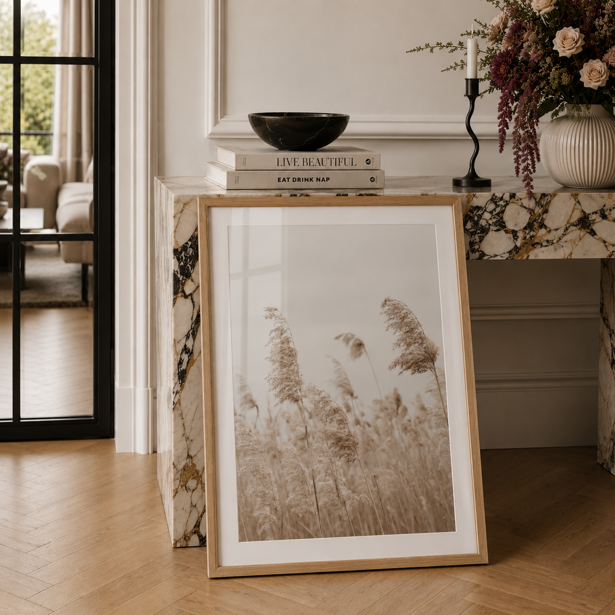 A framed art print featuring a sepia-toned photograph of tall grasses, styled leaning against a marble side table in a bright, modern living room.
