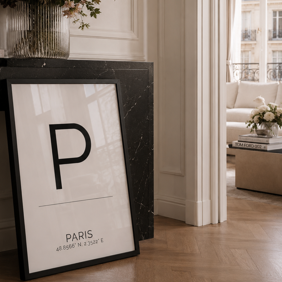 A framed minimalist art print featuring a large black letter 'P' and the text 'PARIS' leans against a black marble console table in a bright, elegant room with a floral arrangement and a window view in the background.