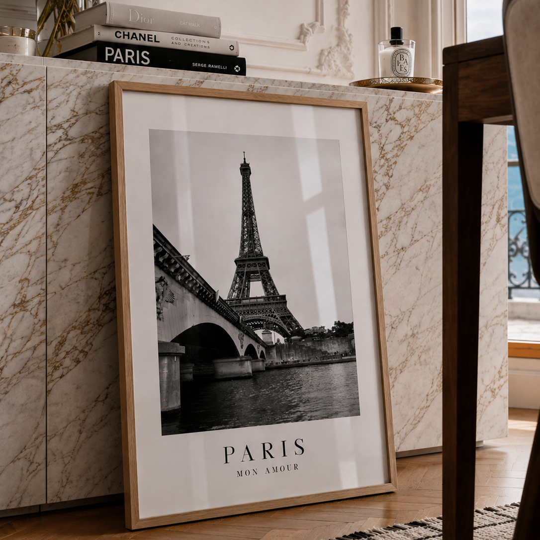 A framed black and white Paris poster leaning against a marble cabinet in a stylishly decorated room. The poster features a black and white photograph of the Eiffel Tower with the text 'PARIS MON AMOUR' at the bottom, enclosed in a light wood frame.