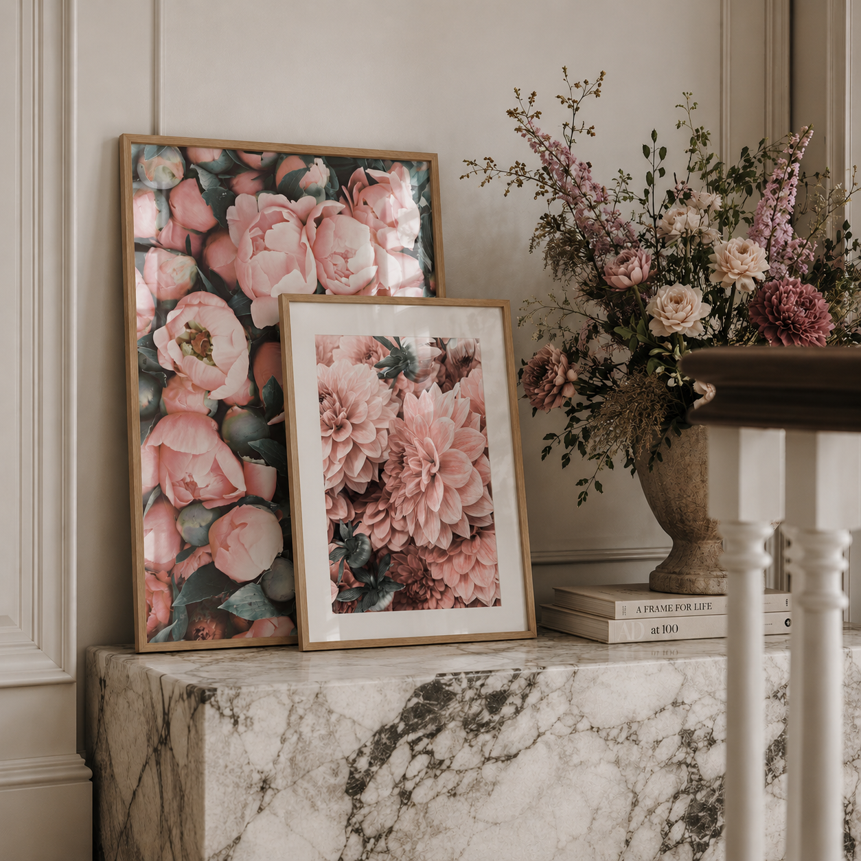 Two framed floral art prints with pink and peach blossoms are displayed on a white marble console table next to a large vase of dried and fresh flowers in a neutral room setting.