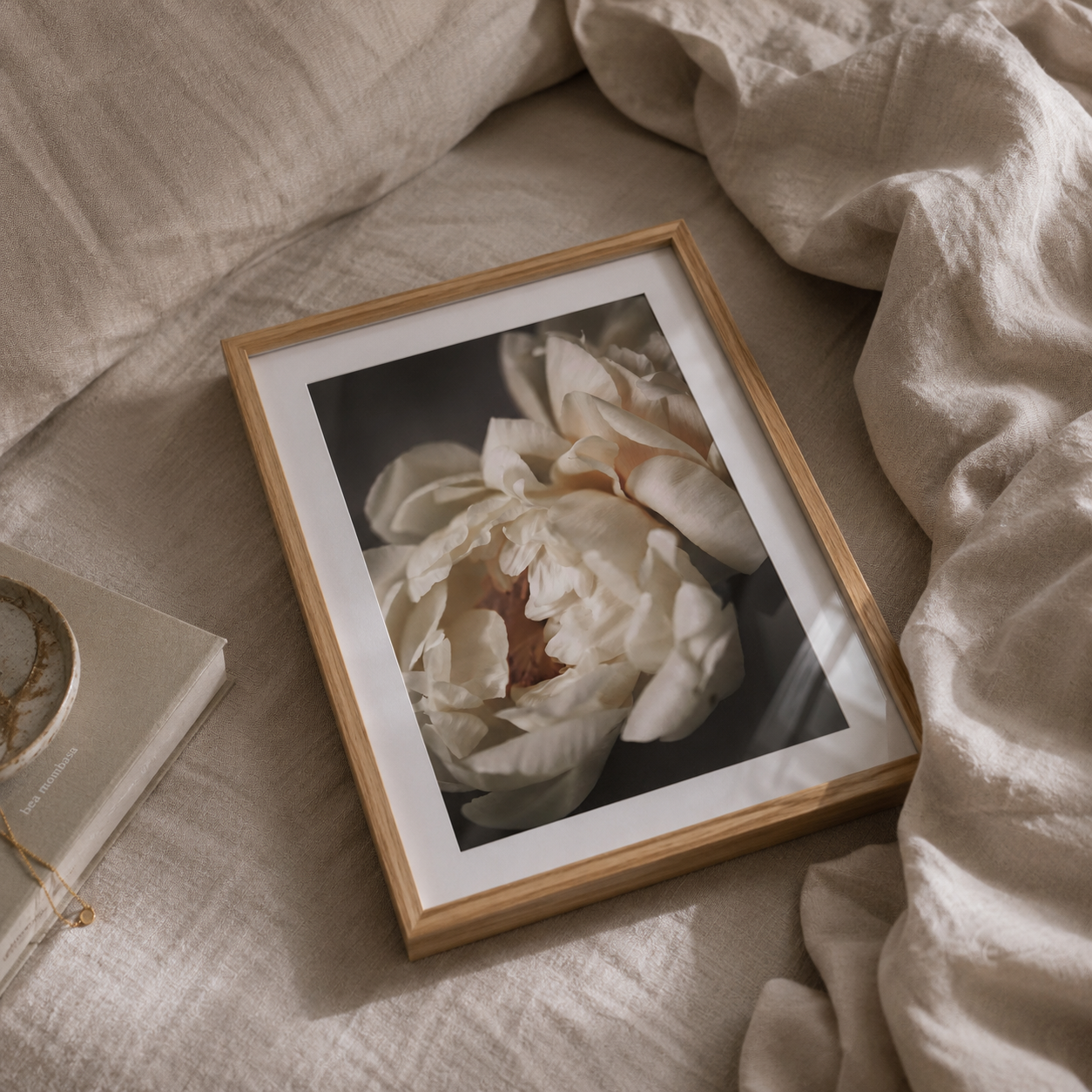 A framed floral art print featuring cream-colored peony blossoms, styled on a beige linen surface with a book and draped fabric nearby.