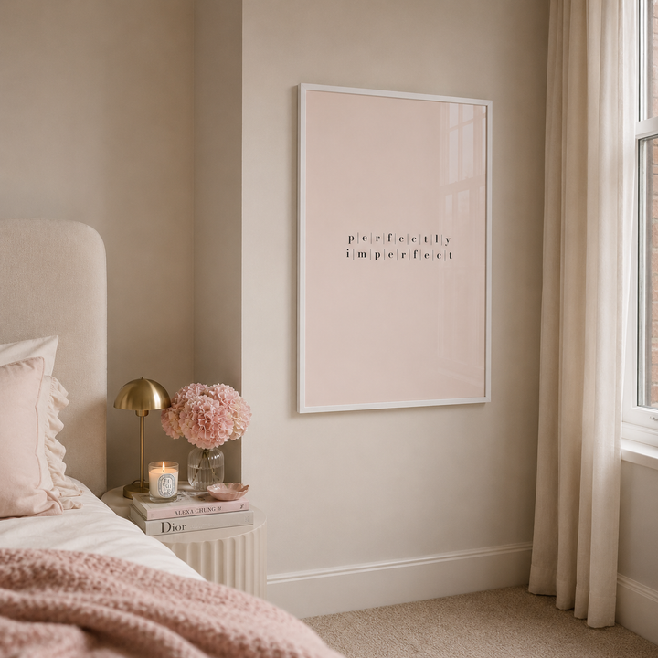 A minimalist framed wall art print with the text 'perfectly imperfect' in black lettering on a pale blush background, styled in a soft beige bedroom next to a bed and a window.