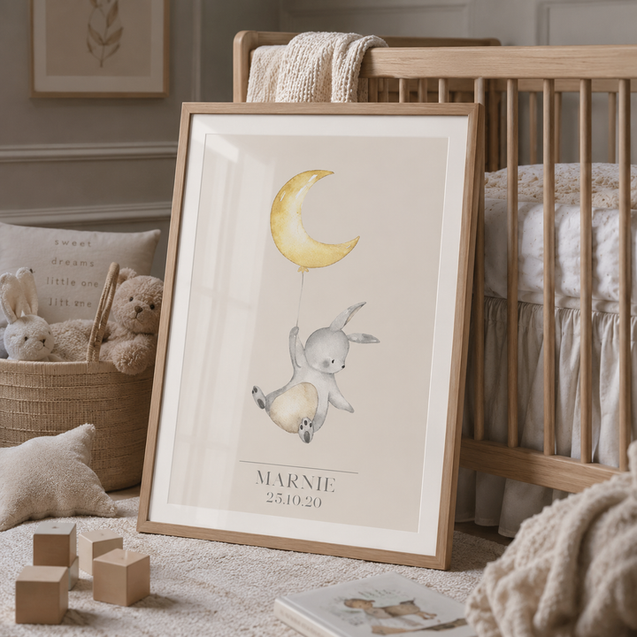 A personalized framed art print featuring a watercolor illustration of a bunny holding a crescent moon, leaning against a wooden crib in a styled nursery setting.