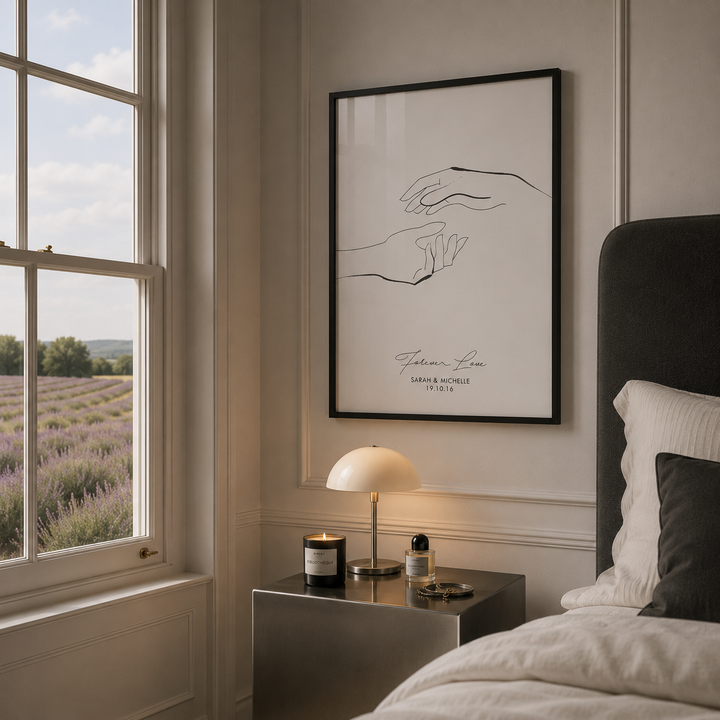 A framed minimalist art print featuring a black line drawing of hands displayed on a bedroom wall above a bedside table with a lamp and candle.