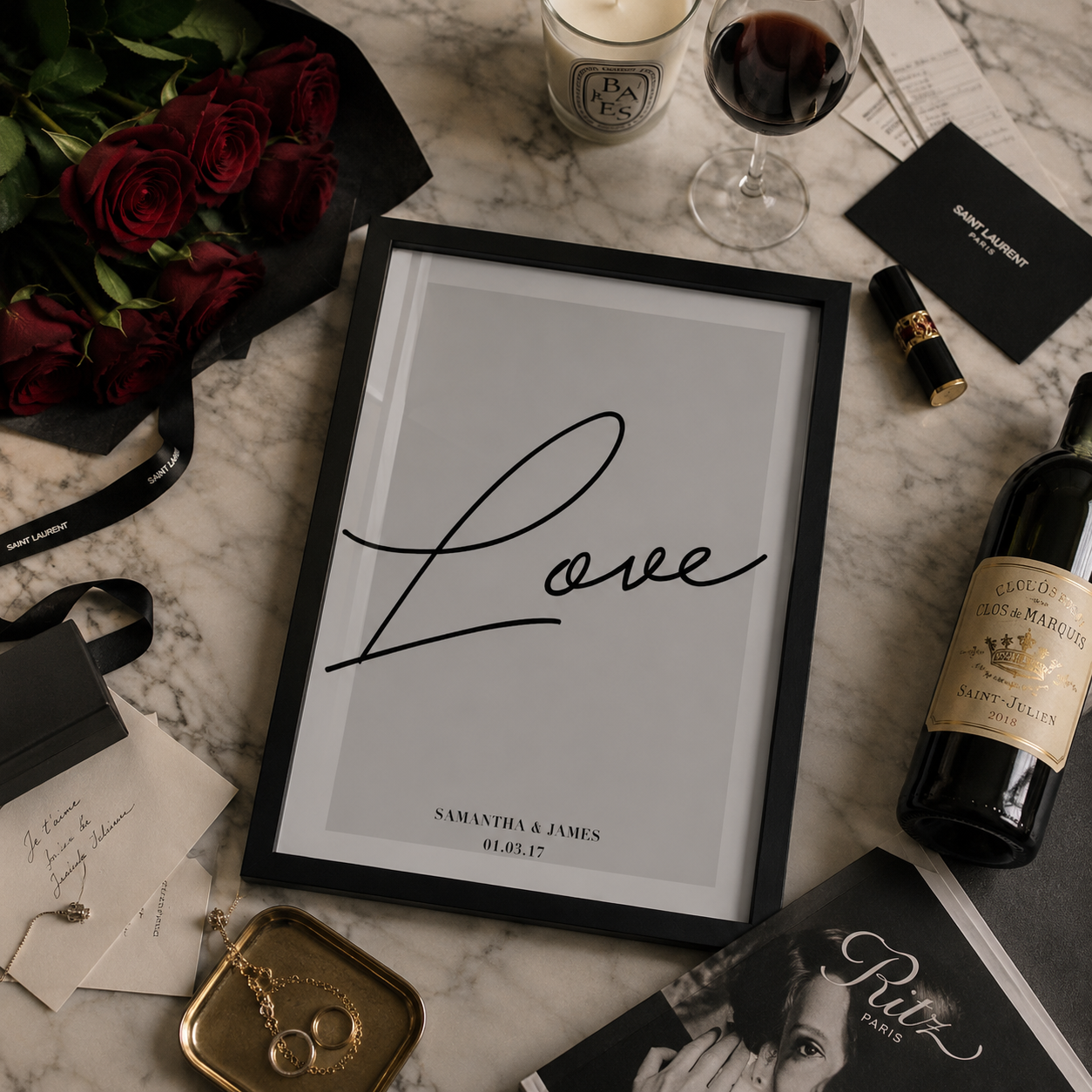 A framed custom wedding art print featuring the word 'Love' in elegant black script on a light gray background, styled in a flat lay with red roses, a glass of red wine, a candle, and other romantic props on a marble surface.