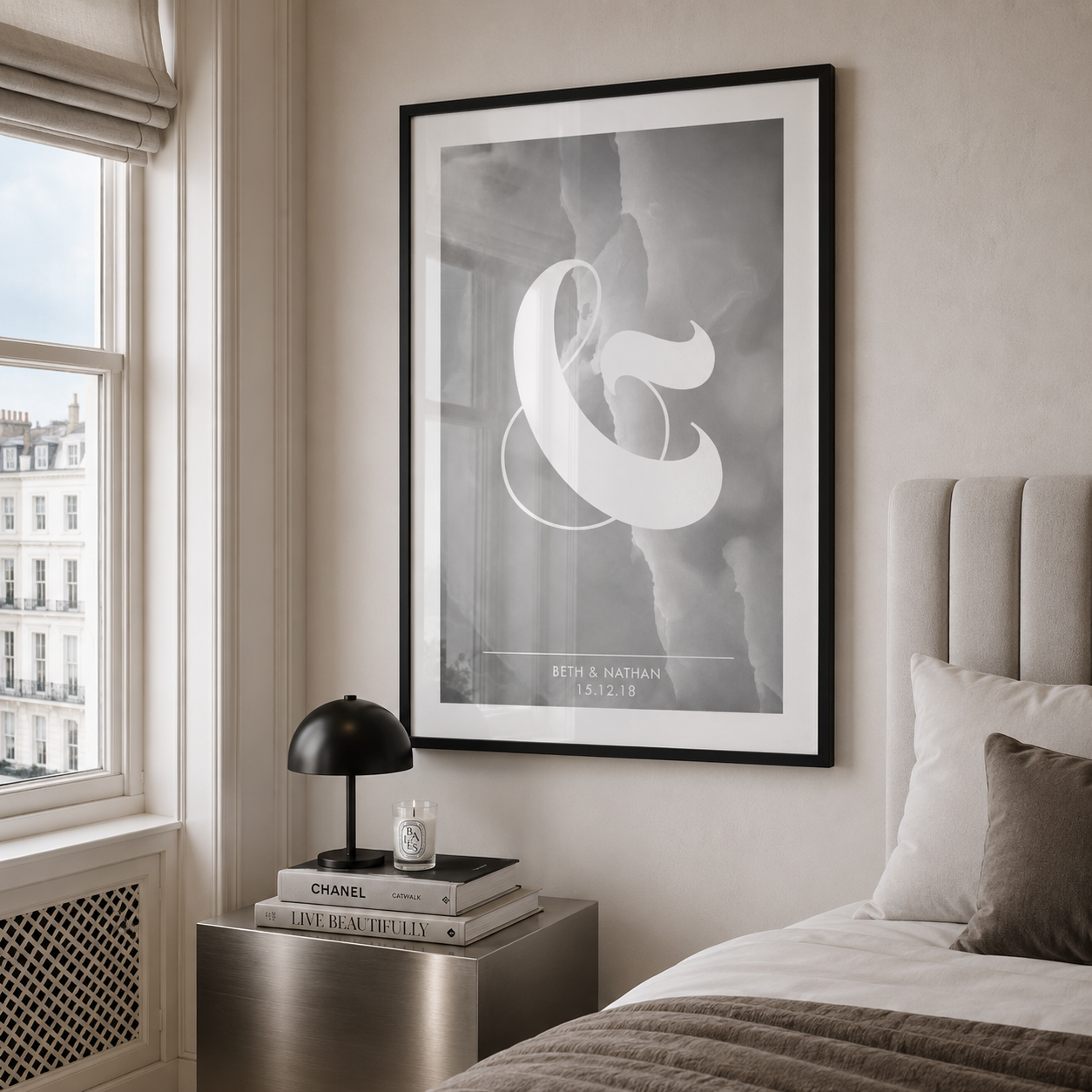 A framed black and white art print featuring a large white ampersand symbol, displayed on a beige wall in a modern bedroom setting next to a window and bedside table.