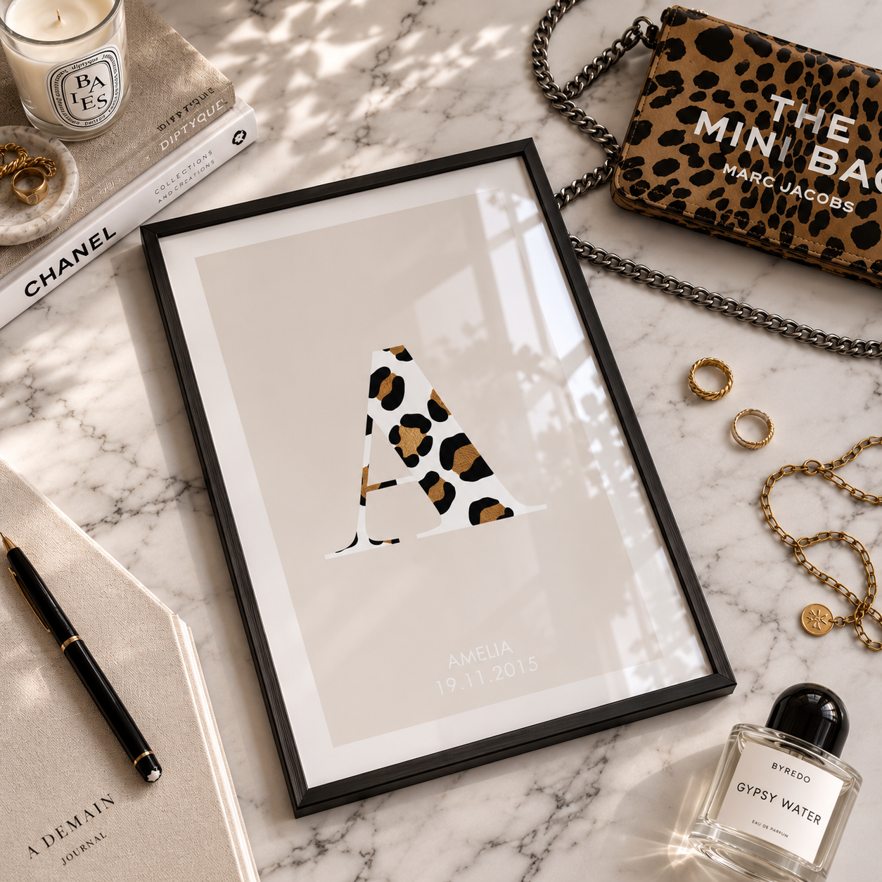 A framed minimalist art print featuring a stylized letter 'A' in a leopard print pattern, styled in a flat-lay on a white marble surface with various fashion accessories and books.