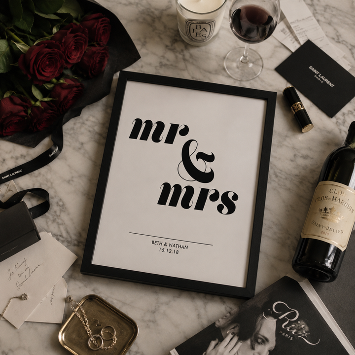 A framed personalized wedding sign reading 'mr & Mrs' with names and date below, styled on a marble surface with roses, wine, candles, rings, and other romantic props.