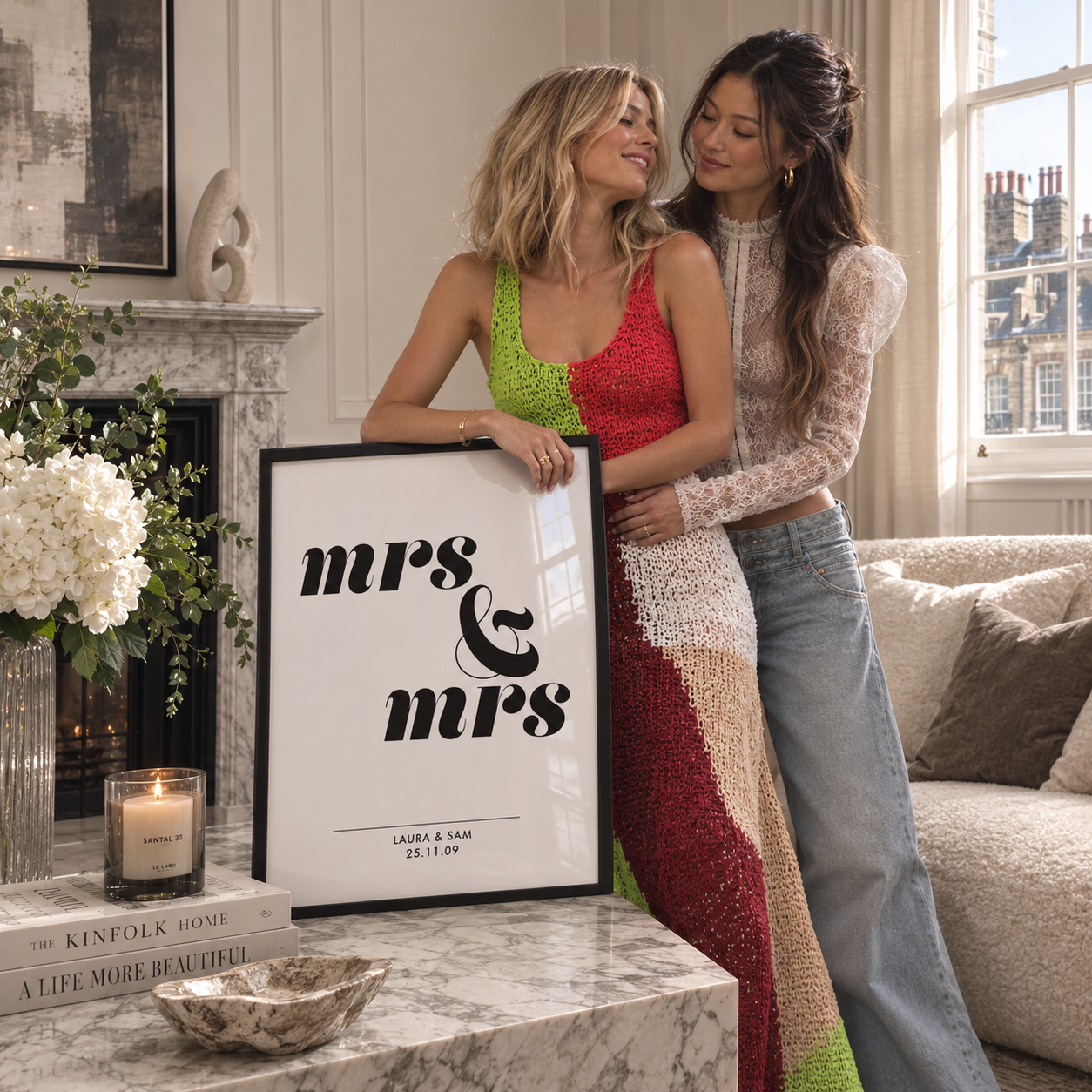 Two women standing indoors beside a framed print that reads 'mrs & mrs' with names and a date below. The room has a fireplace, sofa, flowers, and a candle, so this is a Lifestyle image rather than a clean Product/Packaging shot.