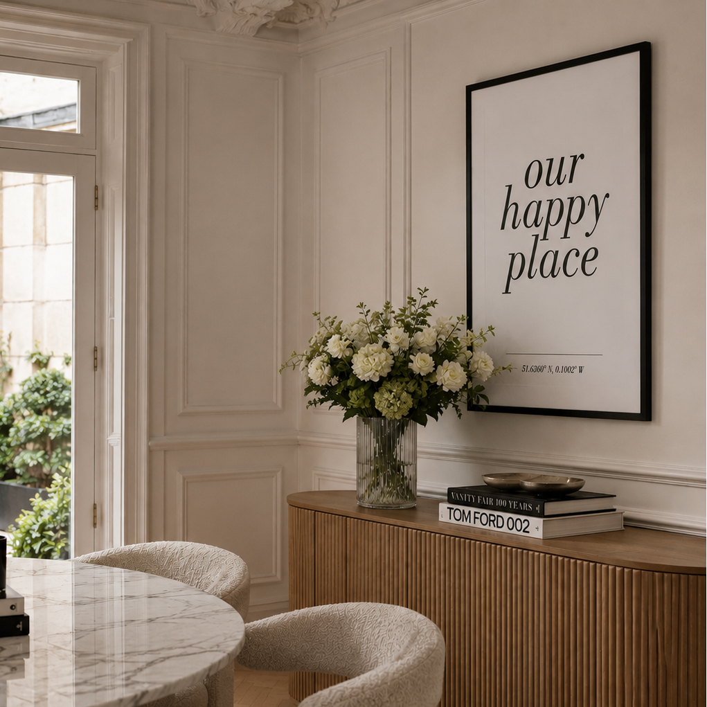 A styled interior room scene with a round marble dining table, textured cream bouclé chairs, a wooden sideboard, framed wall art reading 'our happy place', and a vase of white flowers.