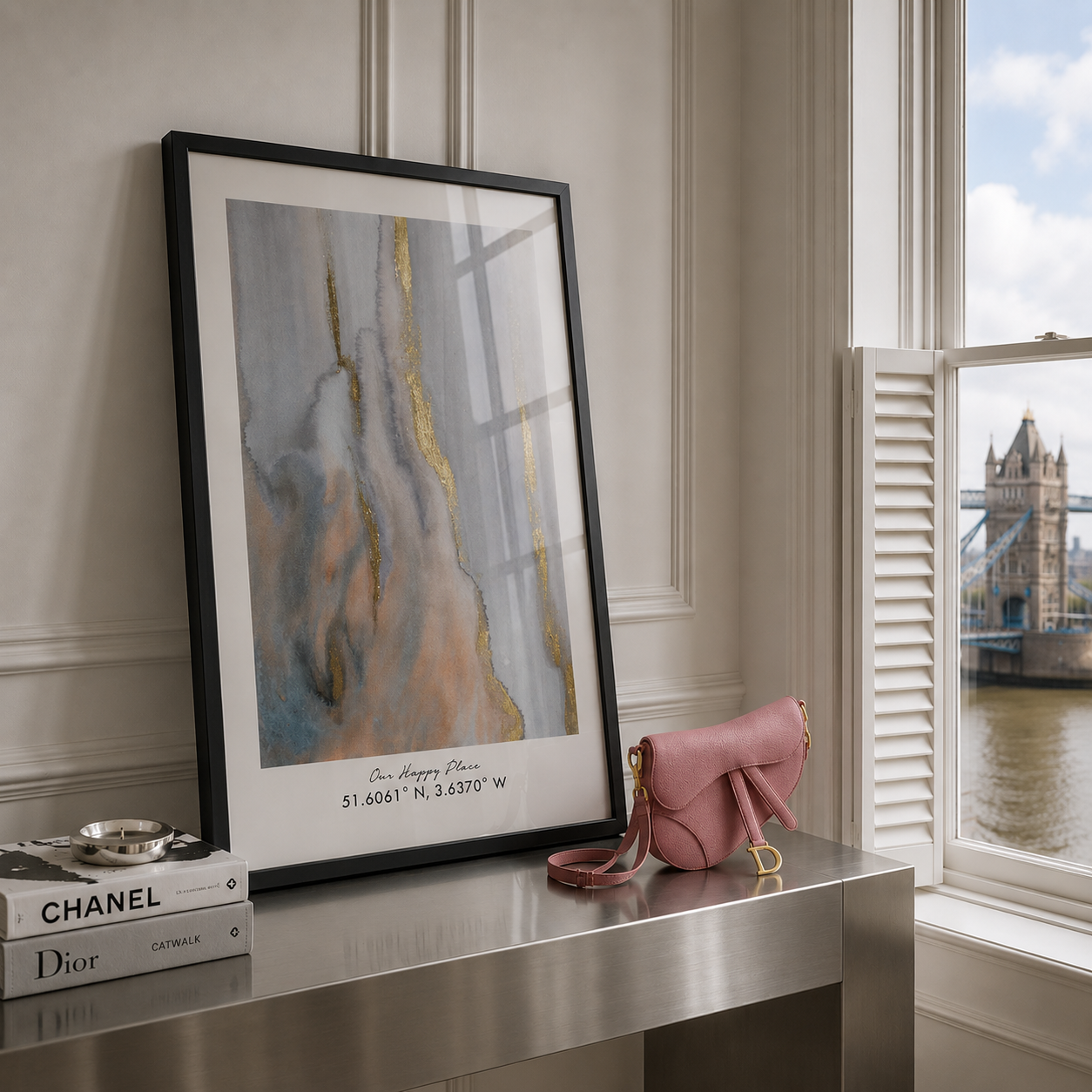 A framed abstract art print with gold and blue-gray marble-like veining leans on a metallic console table beside a pink Dior handbag and books, with a window showing Tower Bridge and the river in the background.