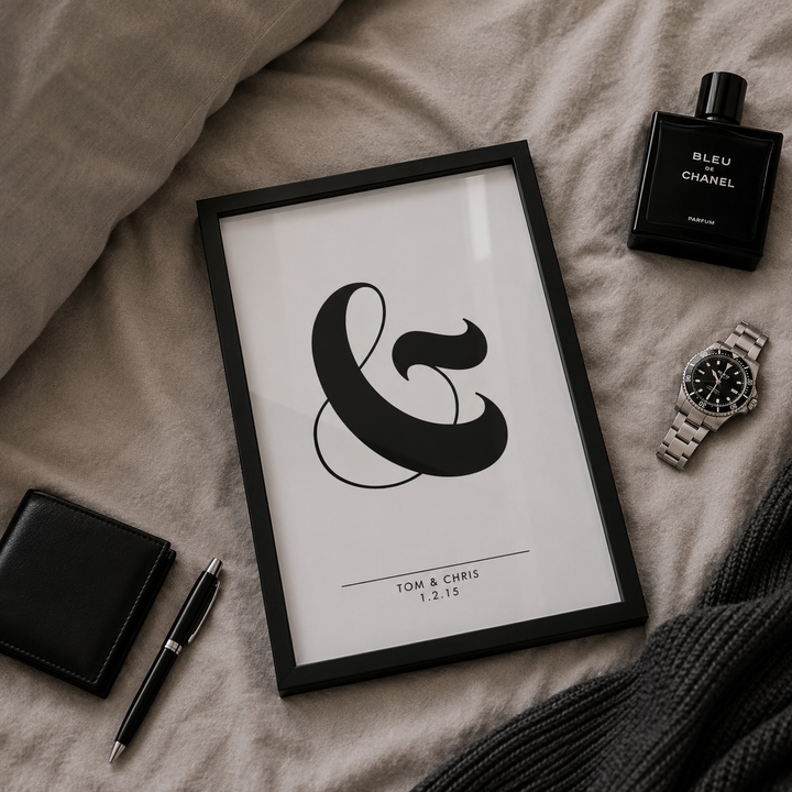 A framed minimalist art print featuring a large black ampersand on a white background, styled in a flat-lay arrangement on beige linen bedding alongside a Chanel perfume bottle, a silver watch, a black wallet, a black pen, and a dark gray knit blanket.
