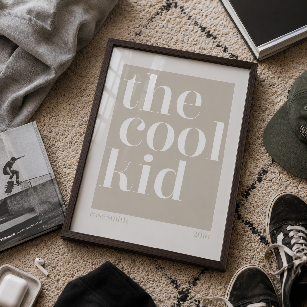 A framed typographic art print reading 'the cool kid' with 'rose smith 2016' at the bottom, styled on a textured beige rug with a gray hoodie, magazine, cap, sneakers, and other casual props around it.
