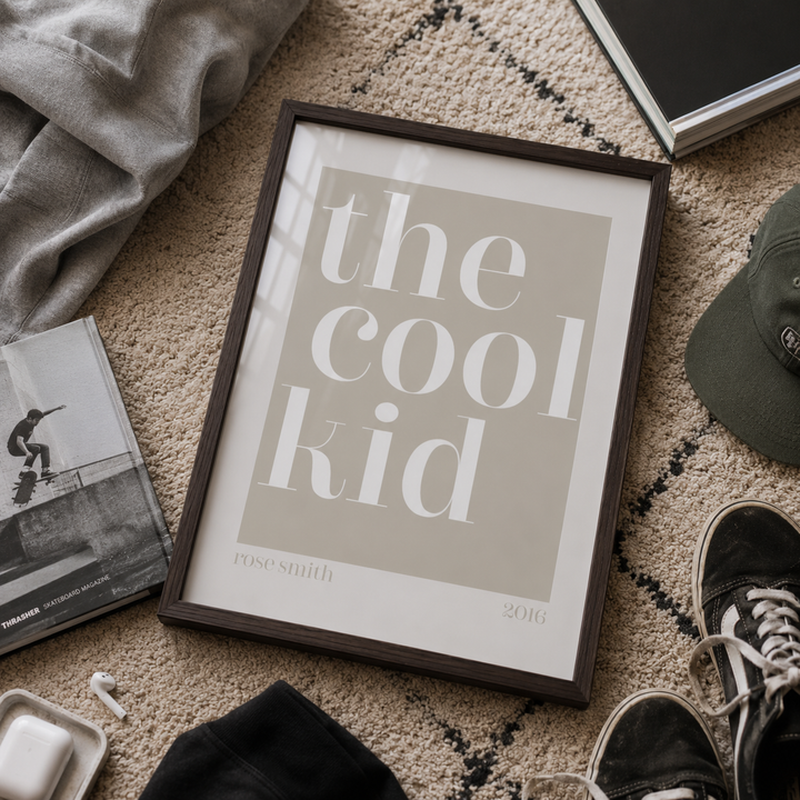 A framed typographic art print reading 'the cool kid' with 'rose smith 2016' at the bottom, styled on a textured beige rug with a gray hoodie, magazine, cap, sneakers, and other casual props around it.