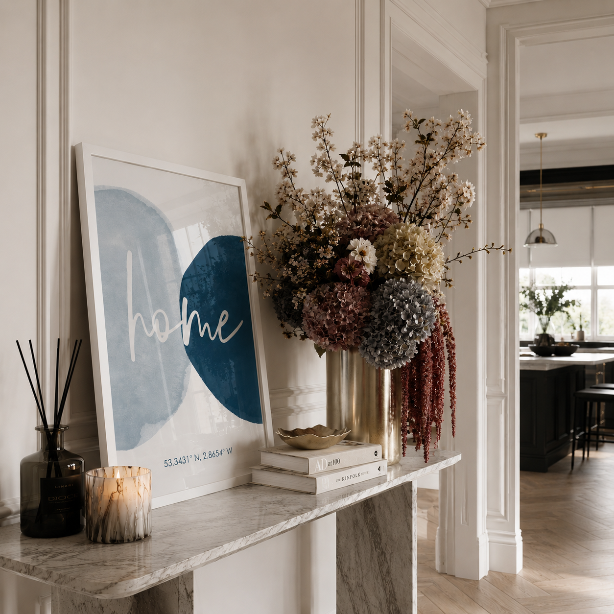A framed abstract art print with the word 'home' in white script over blue and gray shapes, styled on a marble console table in a bright, modern interior next to a dried floral arrangement in a gold vase.