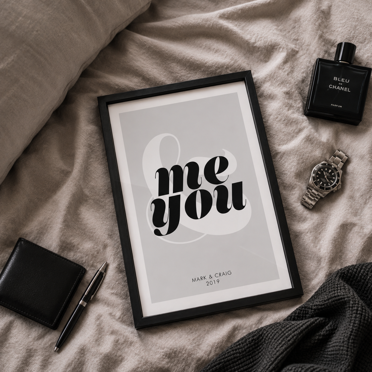 A framed typographic wall art print reading 'me you' with a large ampersand, styled on beige bedding with a black wallet, pen, perfume bottle, watch, and dark knit fabric nearby.
