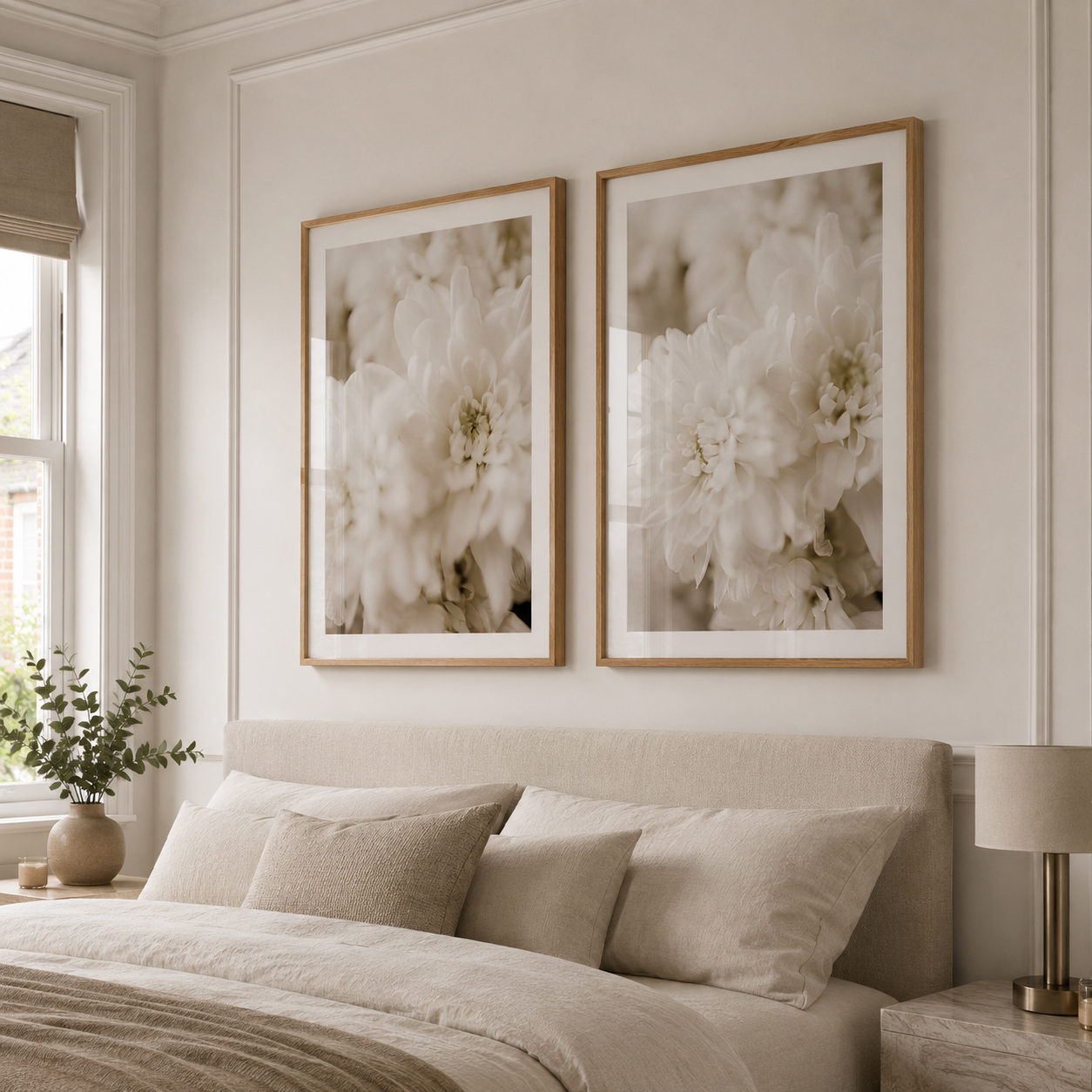 Two framed floral wall art prints with light wood frames hang above a bed in a softly lit neutral bedroom. The artwork shows large white flowers in soft beige and cream tones.