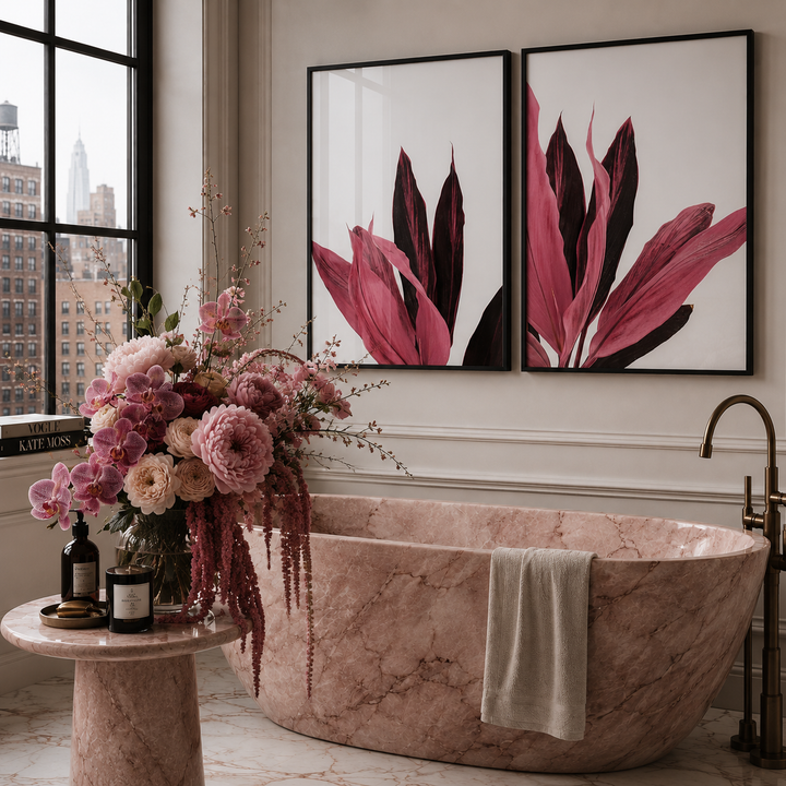 A luxury bathroom scene featuring a freestanding pink marble tub, a matching marble side table with a large pink and purple floral arrangement, and two framed botanical wall art prints above the tub.