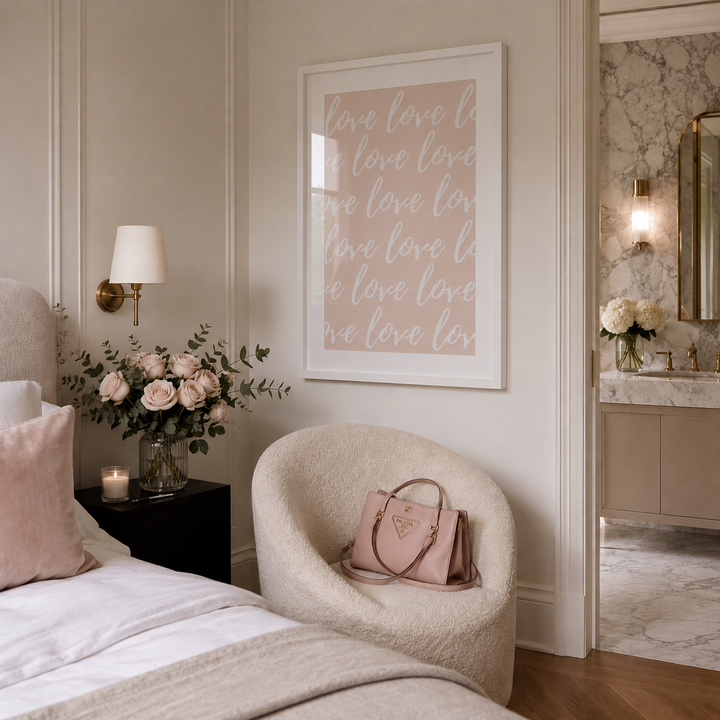 A framed vertical art print with the word 'love' repeated in white script on a blush pink background, hanging on a beige wall in a styled bedroom. The room includes a cream textured armchair with a pink handbag, a bed with white linens and a pink pillow, a black nightstand with a flower arrangement, and a doorway leading to a marble bathroom.