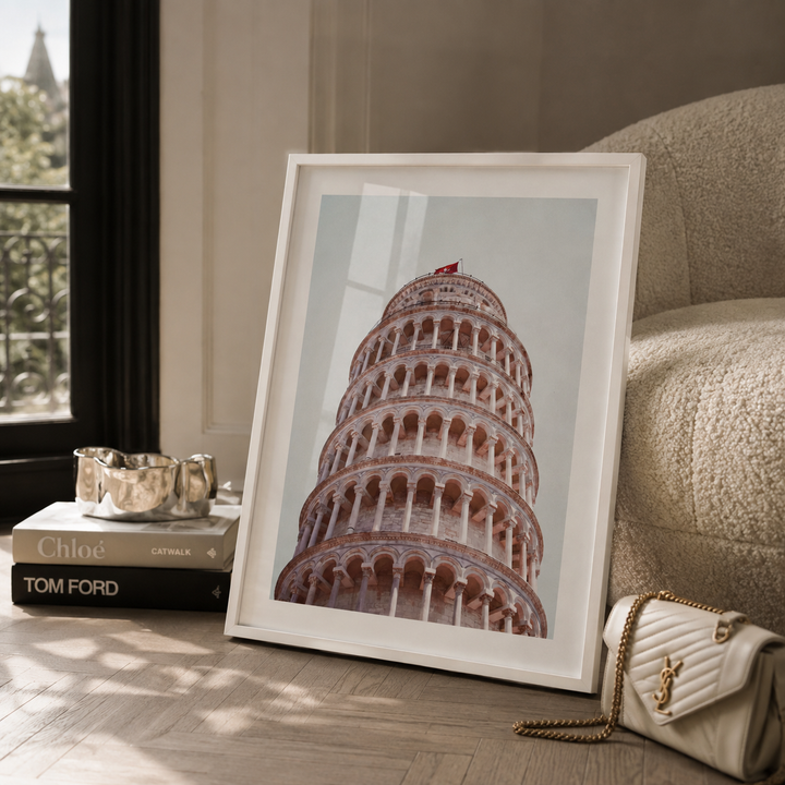 A white-framed art print of the Leaning Tower of Pisa leaning against a beige armchair on a wooden floor, styled with books, a silver bowl, and a white quilted handbag.