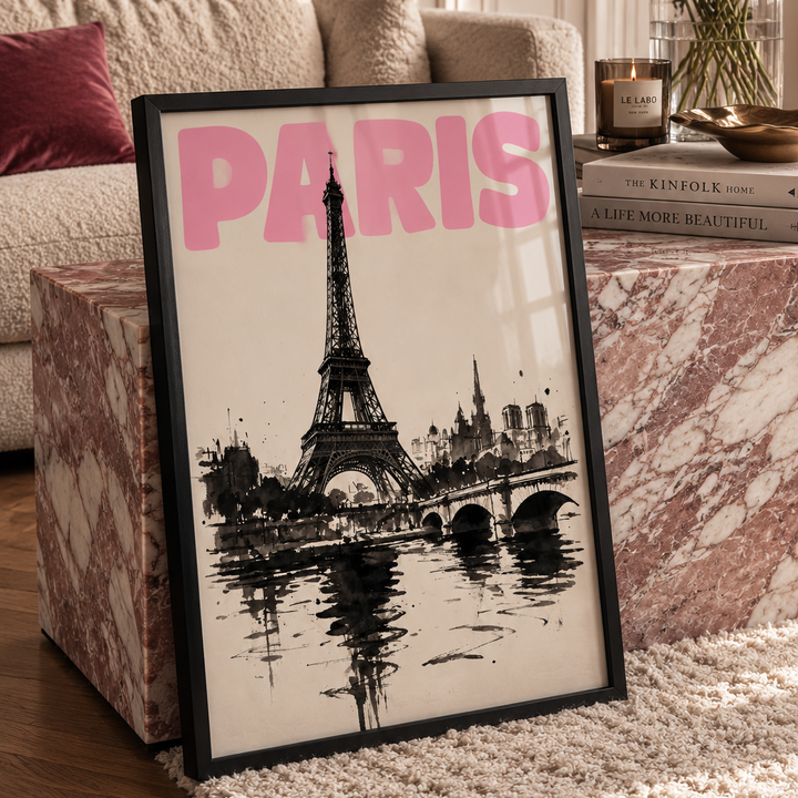 A framed black and white Paris cityscape print with bold pink 'PARIS' lettering, leaning on a marble side table in a styled living room with flowers and a sofa in the background.