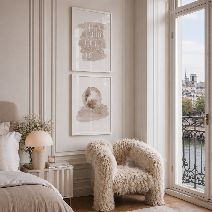 A styled bedroom corner with two framed abstract art prints hanging on a cream paneled wall above a fluffy Freyja Chair by Shropshire Design, next to a bed and a large window overlooking a river and the Paris city skyline.